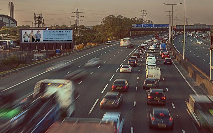 Motorway with a advertising billboard next to it showing Wombats band show.